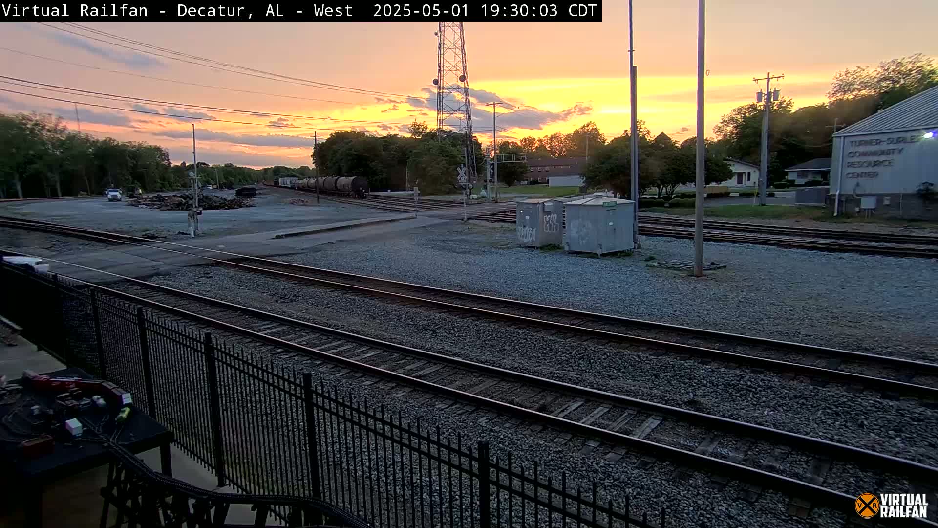 A sunset casts an orange glow over a railroad yard with several train cars, a metal fence, and buildings in the background.