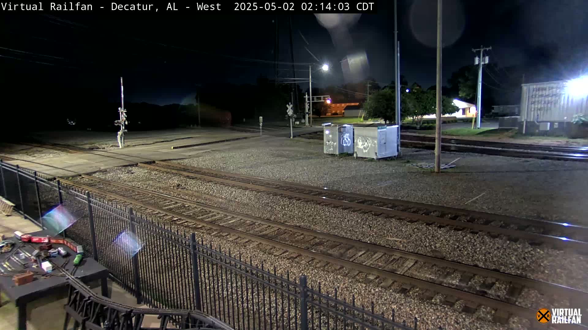 Under a dark night sky, several sets of train tracks and a nearby fenced-in area are visible, along with some utility boxes and a model train setup.