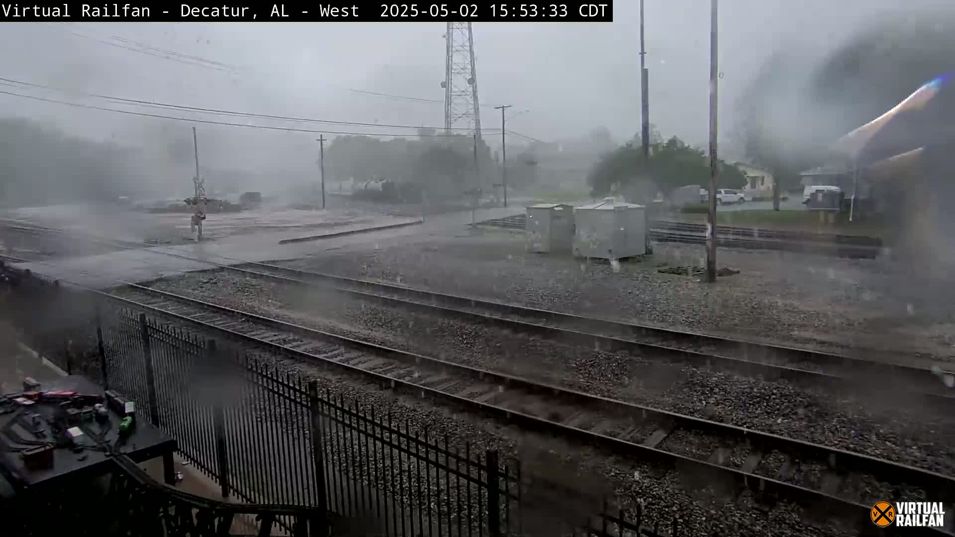 A heavy rainstorm falls on a railway yard, obscuring visibility and showing water collecting on the tracks and surrounding ground.
