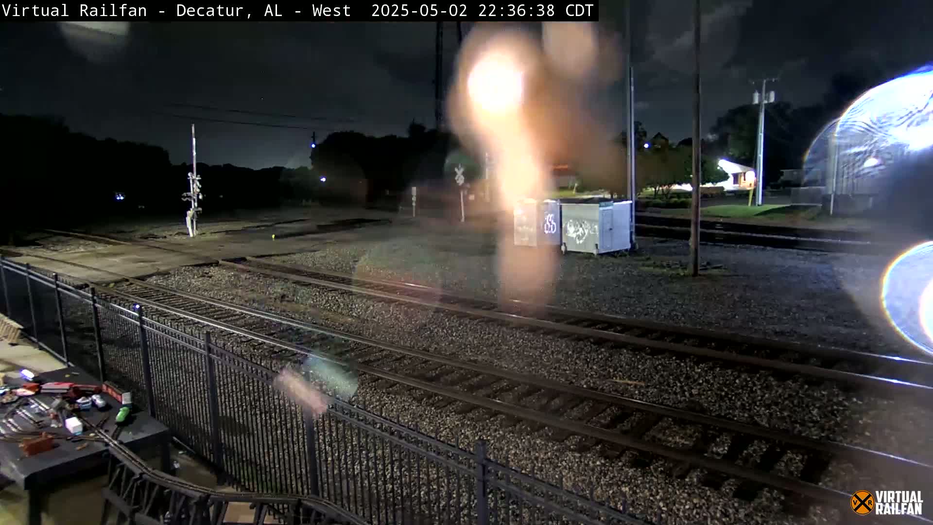 A nighttime view of railroad tracks, a metal fence, and some graffitied dumpsters under a dark, slightly cloudy sky.