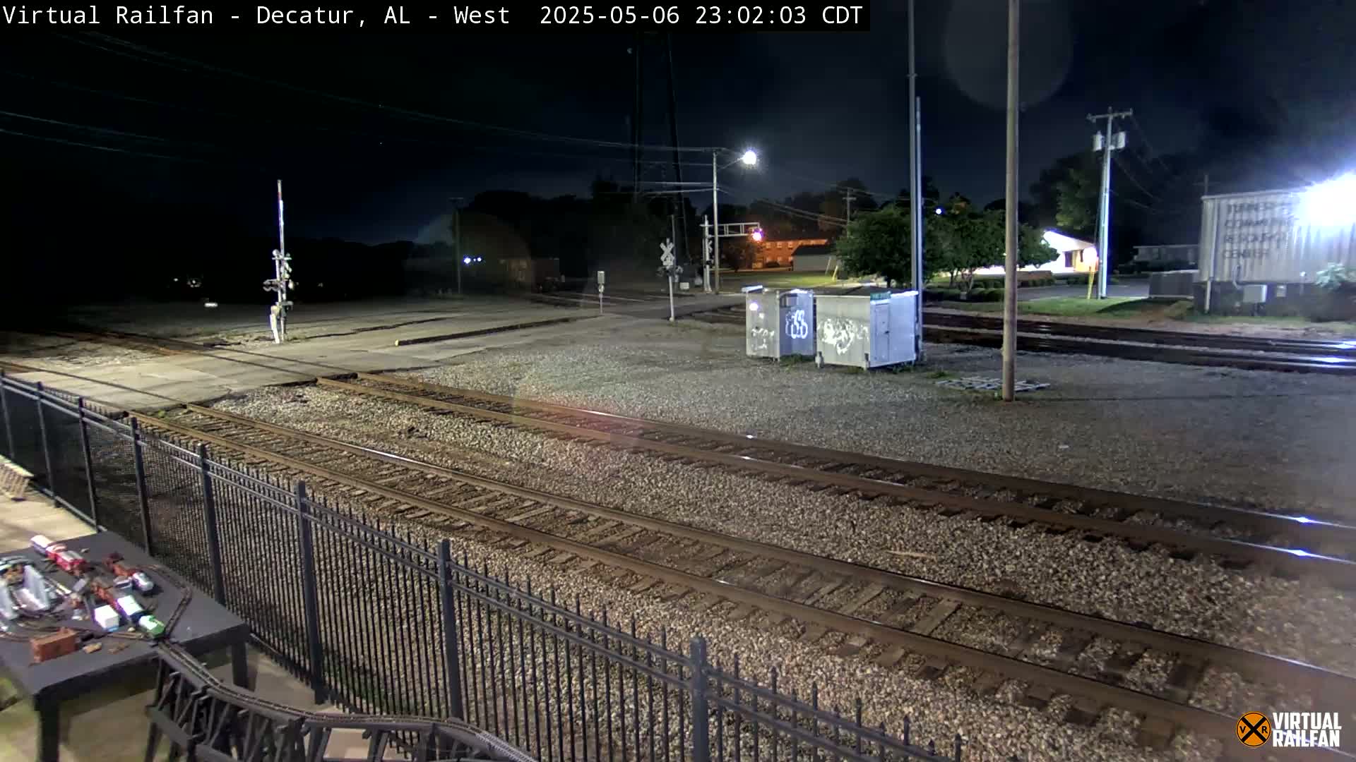 A nighttime view of railroad tracks and switching areas, with some buildings and trees visible in the background under a dark sky.