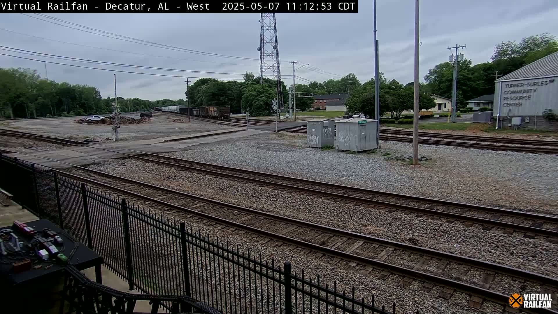 A view of several railroad tracks intersecting, with a gravel surface, a chain-link fence, and some buildings in the background under an overcast sky.