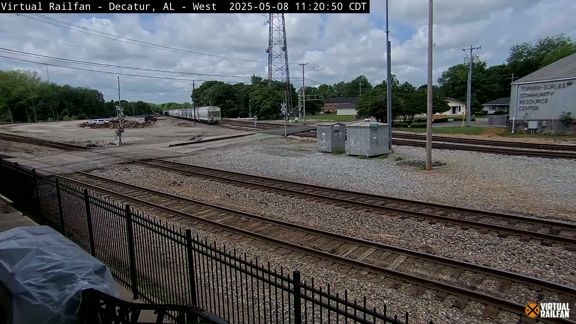 A mostly sunny day shows a railway yard with several train tracks, some rail cars, and a few buildings in the background.