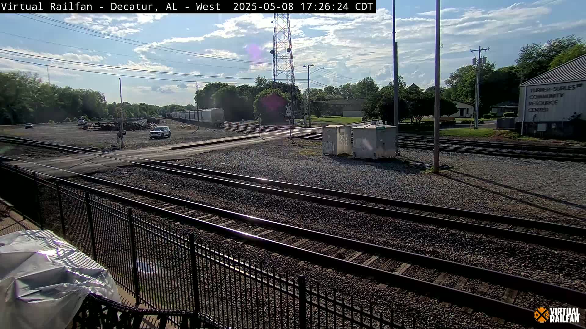 A sunny day reveals a railroad crossing with several train tracks, a roadway, and some vehicles, bordered by trees and a community center.