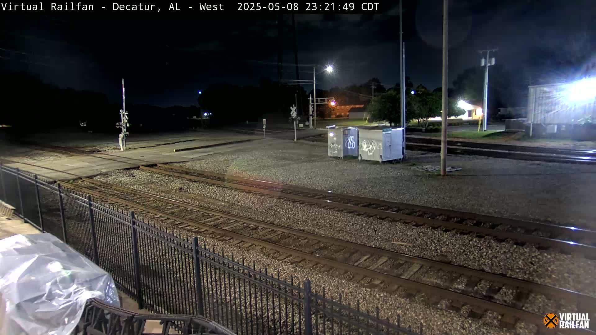 At night, under a clear sky, several sets of train tracks and a metal fence are visible, with some graffiti-covered structures nearby.
