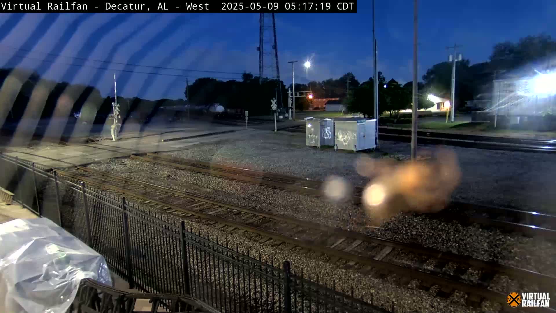 Under a twilight sky, railroad tracks extend into the distance past a metal fence, with some buildings and trees visible in the background.