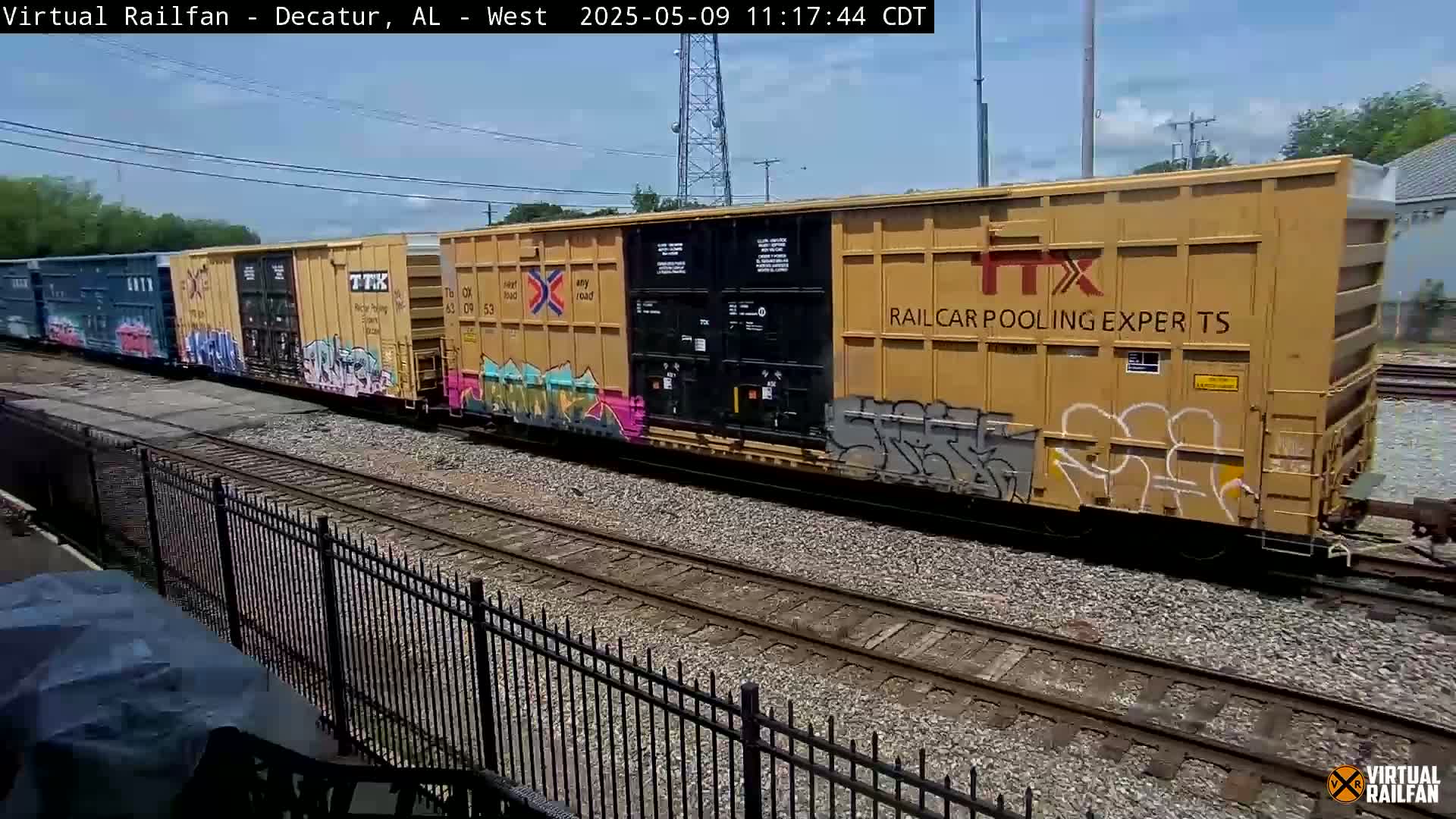 A freight train with graffiti-covered cars moves past a black metal fence on a sunny day.
