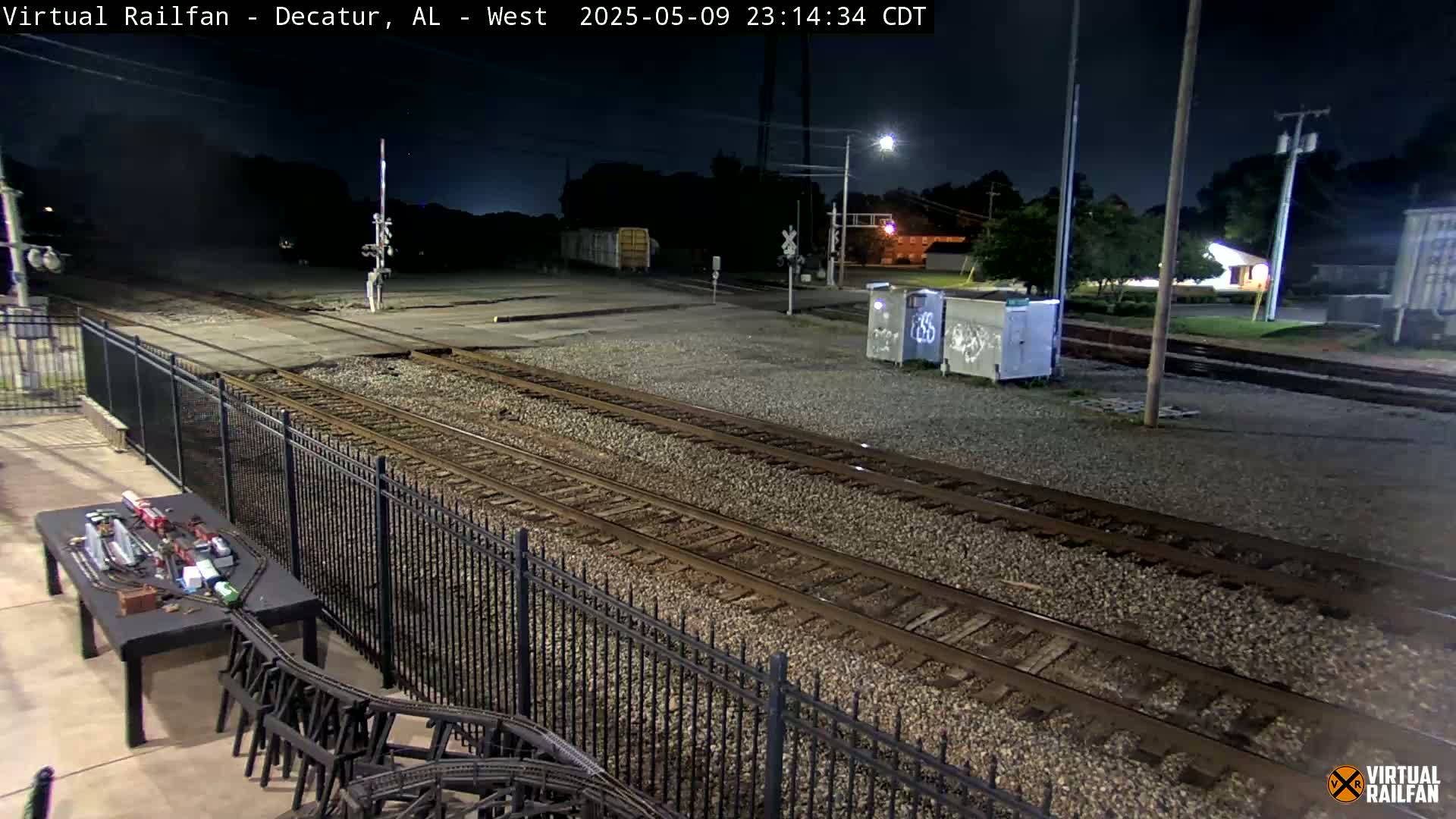 A nighttime view of train tracks, a model train set on a table, and a metal fence, under a clear sky.