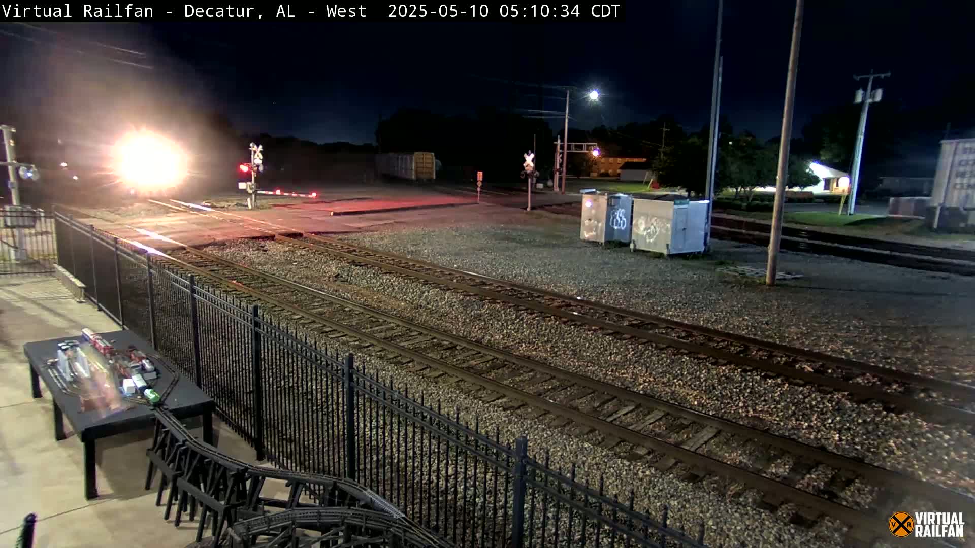 A brightly lit train passes through a nighttime railroad crossing, with a black metal fence and gravel ballast visible.