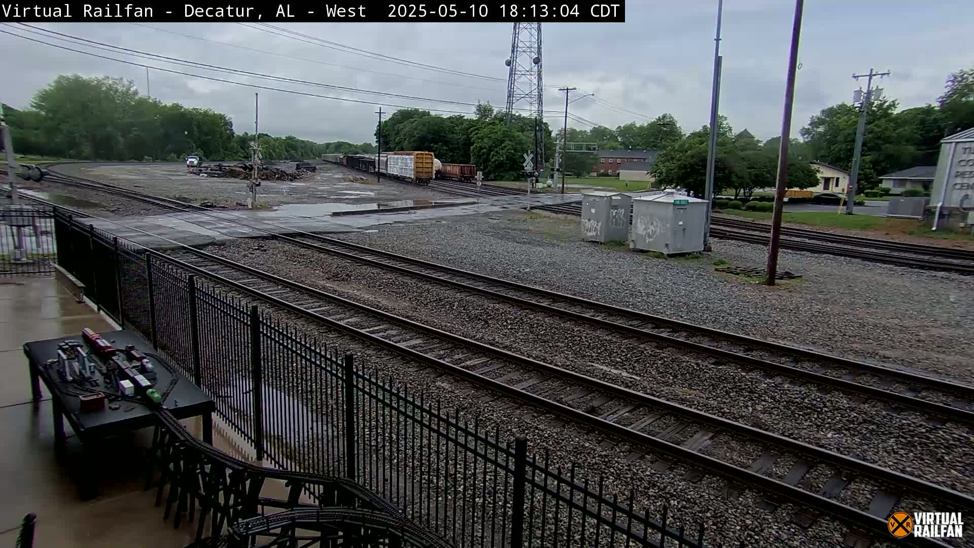 A rainy view of railroad tracks and a train, with some buildings and trees in the background.