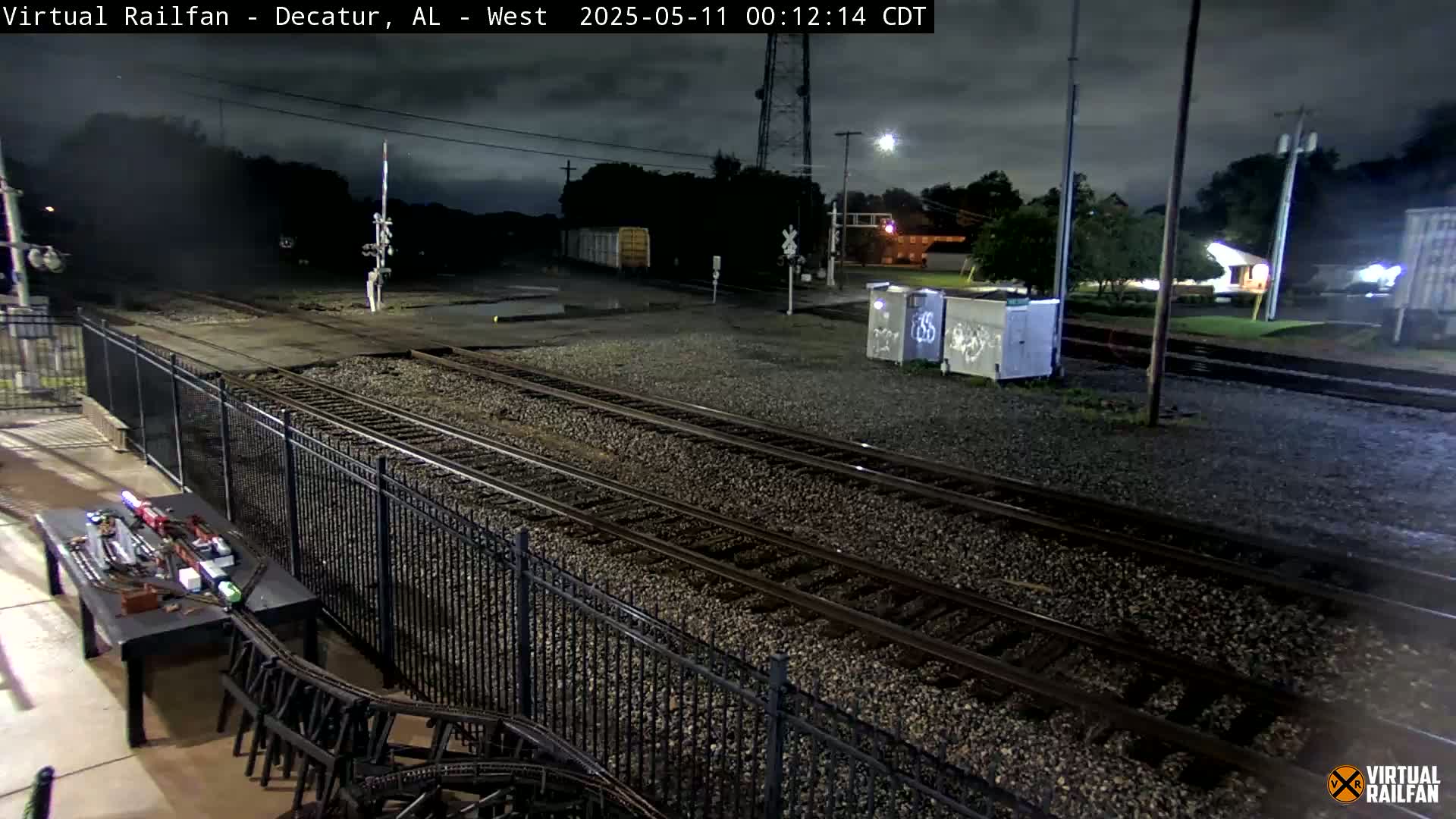 A nighttime view of several train tracks, a metal fence, and a model train set on a table, under a dark, cloudy sky.