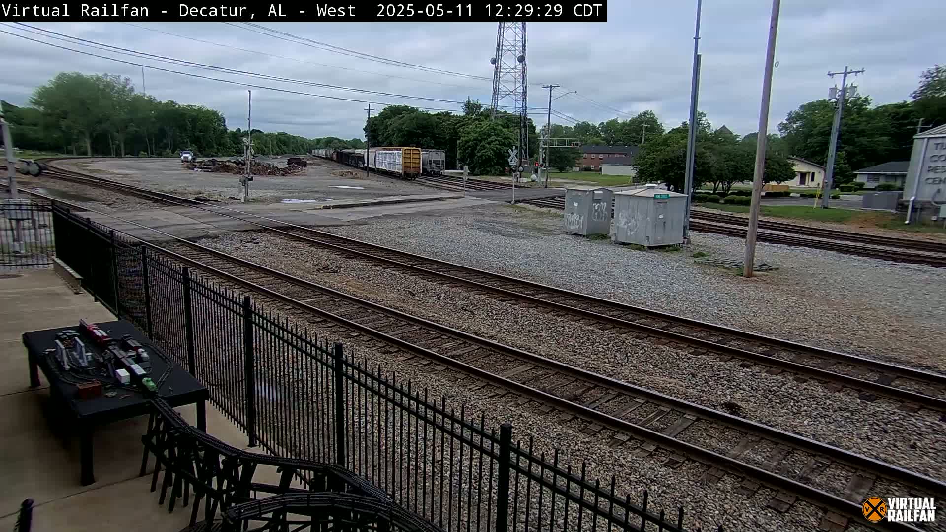 A cloudy day at a train yard shows multiple train tracks, a parked train, and some buildings in the distance.