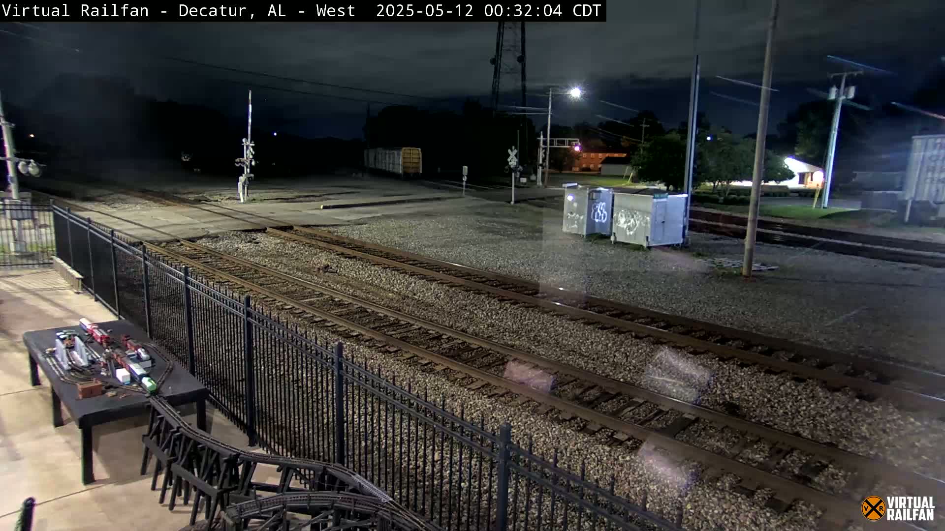 A nighttime view of railroad tracks, bordered by a fence, with a model train set visible on a nearby table under a dark, cloudy sky.