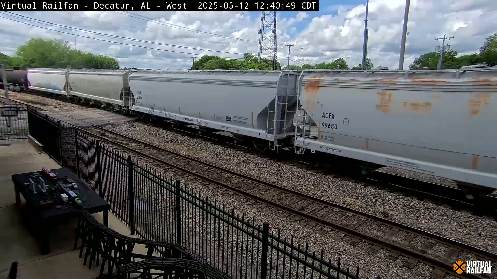 A long freight train of light gray covered hopper cars passes by under a partly cloudy sky.