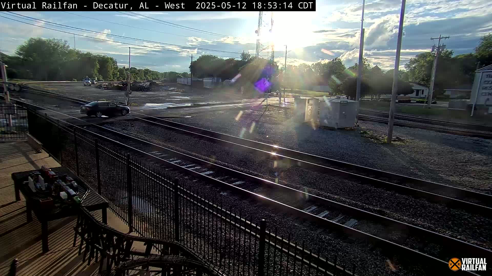 A partly sunny afternoon scene shows multiple train tracks, a car driving across them, and a patio area with outdoor furniture in the foreground.