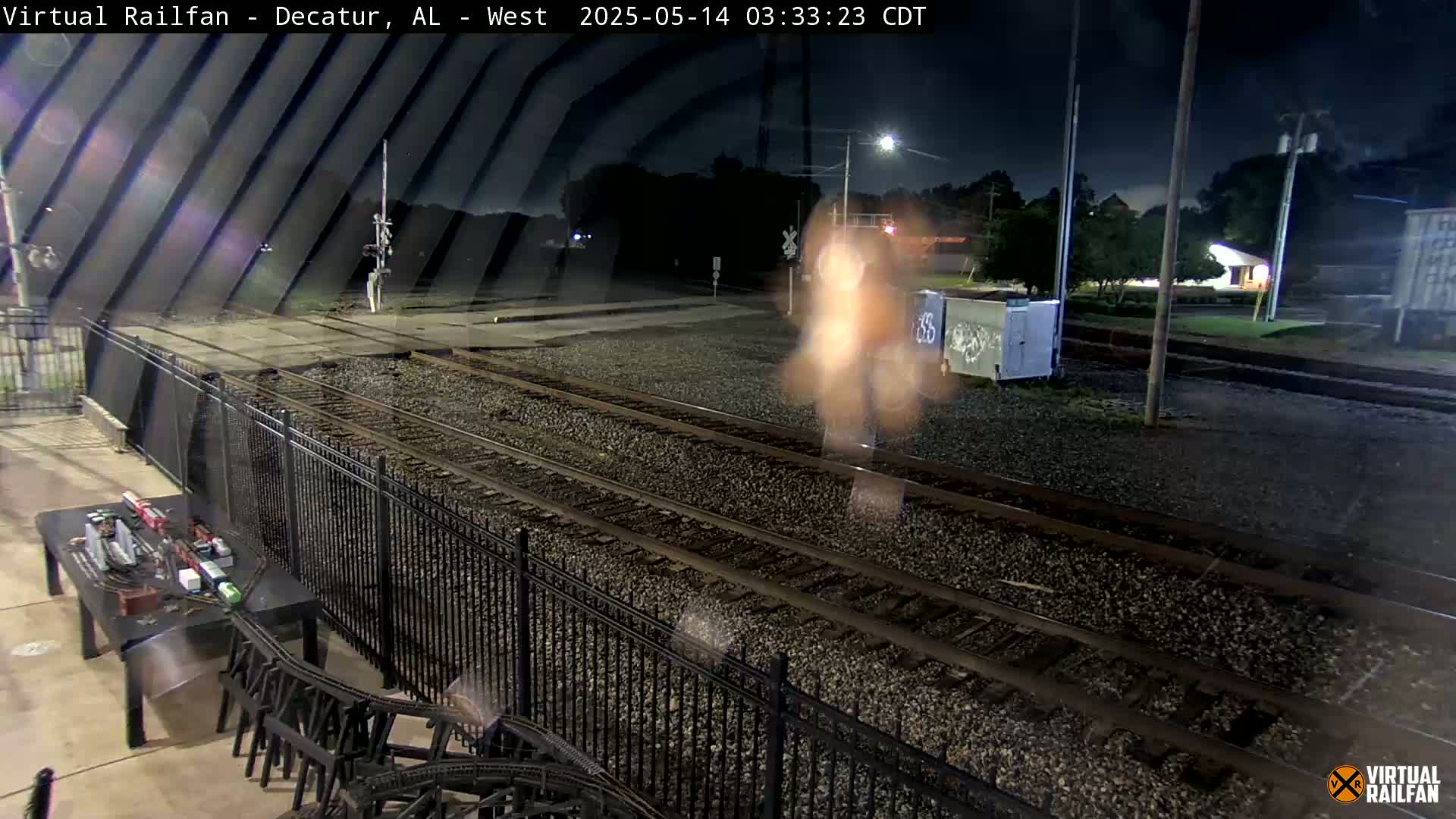 At night, a blurry image shows a train passing by railroad tracks next to a black metal fence, with a model train set visible on a table in the foreground.