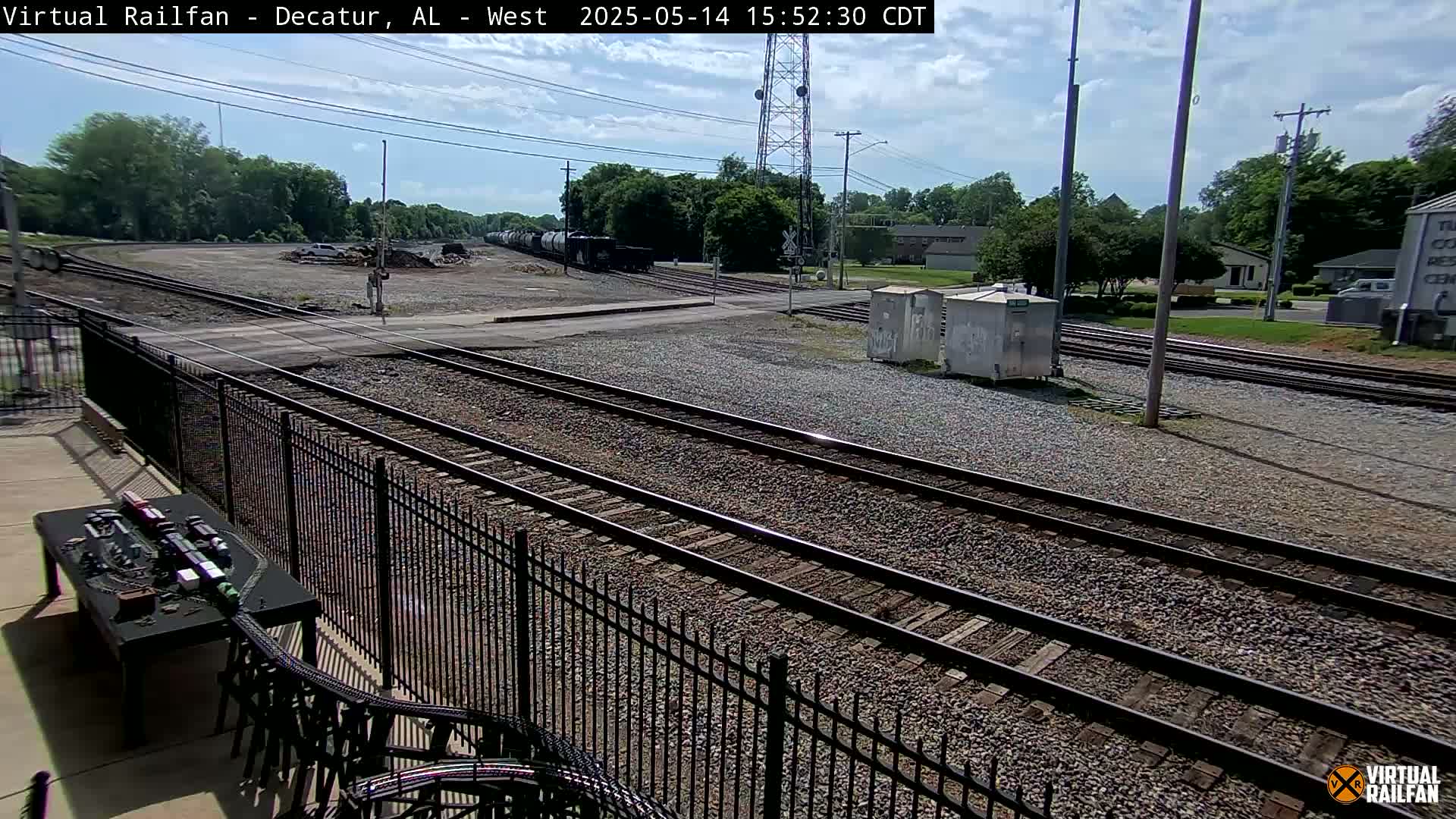 A sunny day view of multiple train tracks intersecting, with a model train set visible in the foreground and several railcars in the background.