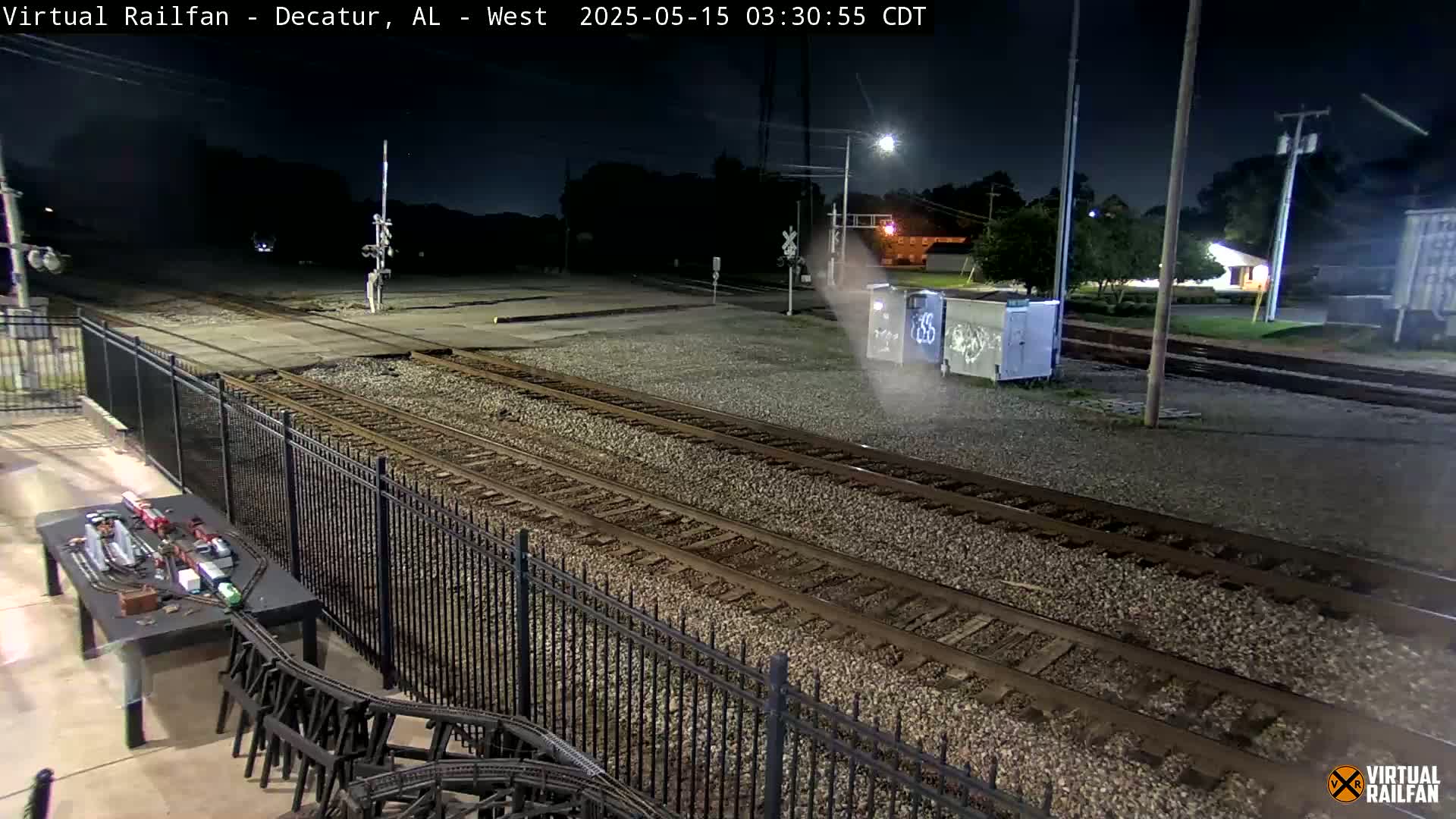 A nighttime view of train tracks, a model train setup, and a black metal fence under a dark sky.