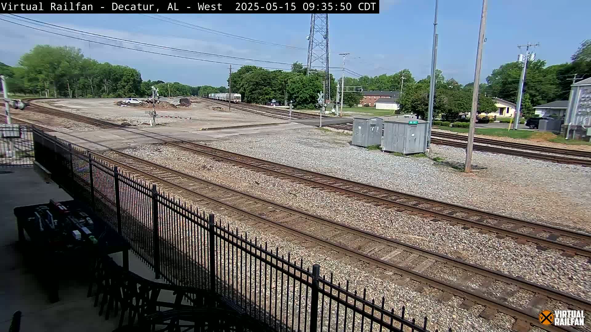 A sunny view of railroad tracks and switching yards, with a black metal fence in the foreground and buildings and trees visible in the background.