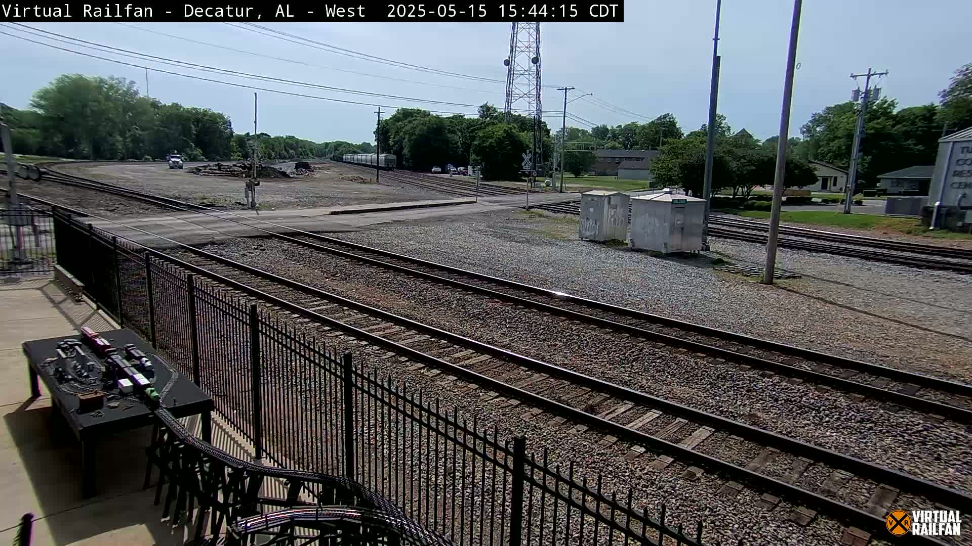 A sunny view of several sets of train tracks, a metal fence, and a model train set on a table, all situated near a roadway and some buildings.