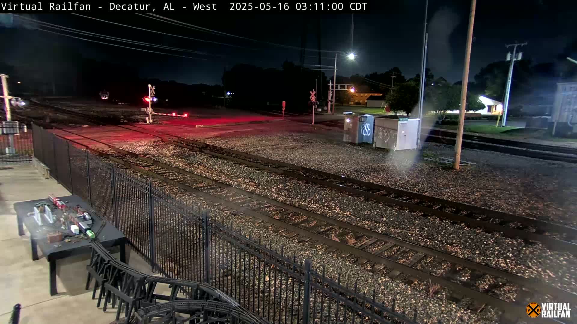 A nighttime view of railroad tracks at a crossing, with a black metal fence separating the tracks from a paved area with a table and chairs.