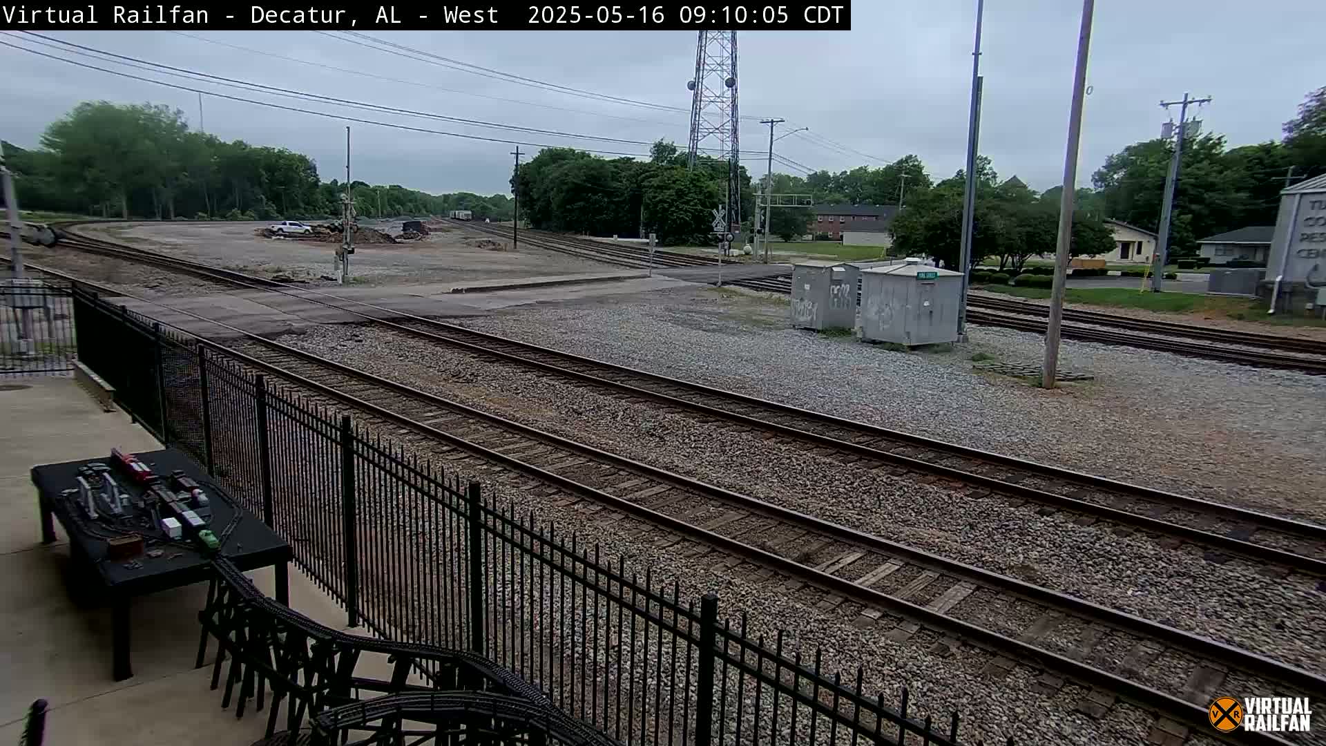 A cloudy day view of multiple train tracks intersecting, with a gravel area and some buildings in the background, next to a black metal fence.