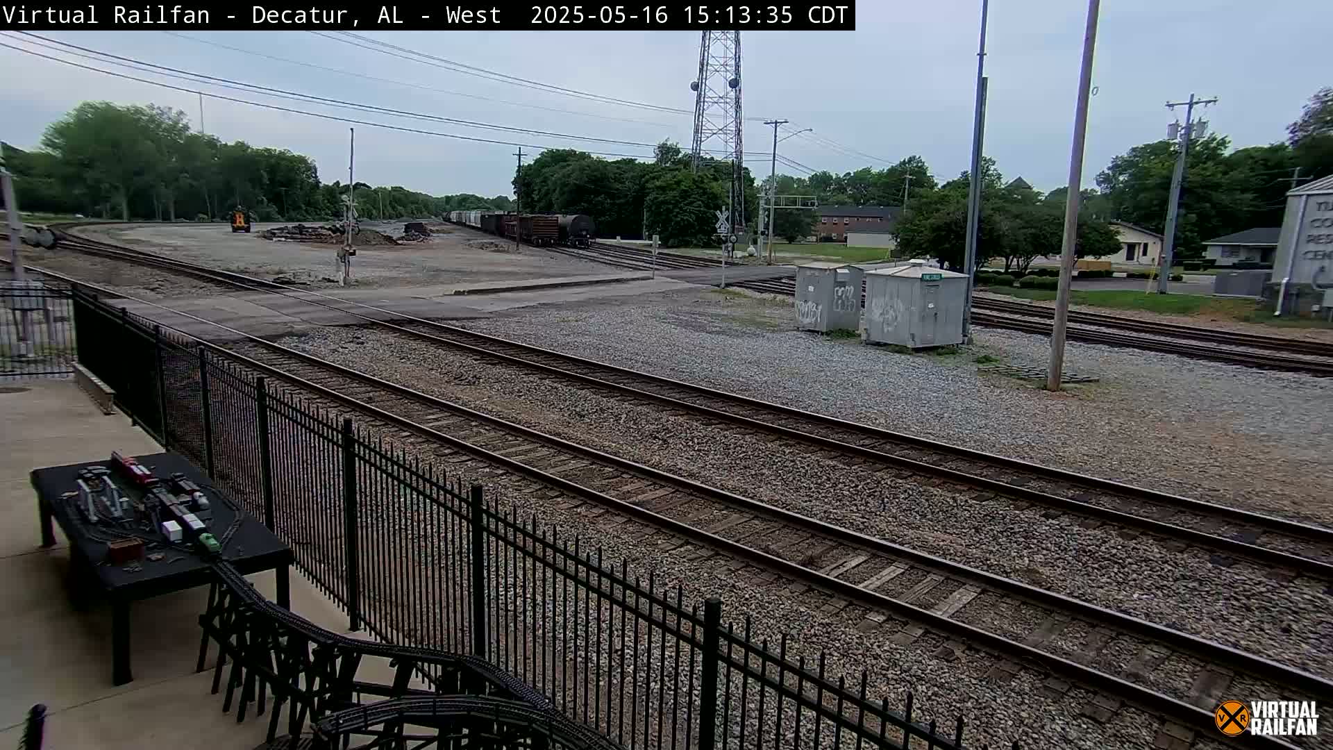 A mostly cloudy day shows several train tracks intersecting near a fenced-in area with some buildings and trees in the background.