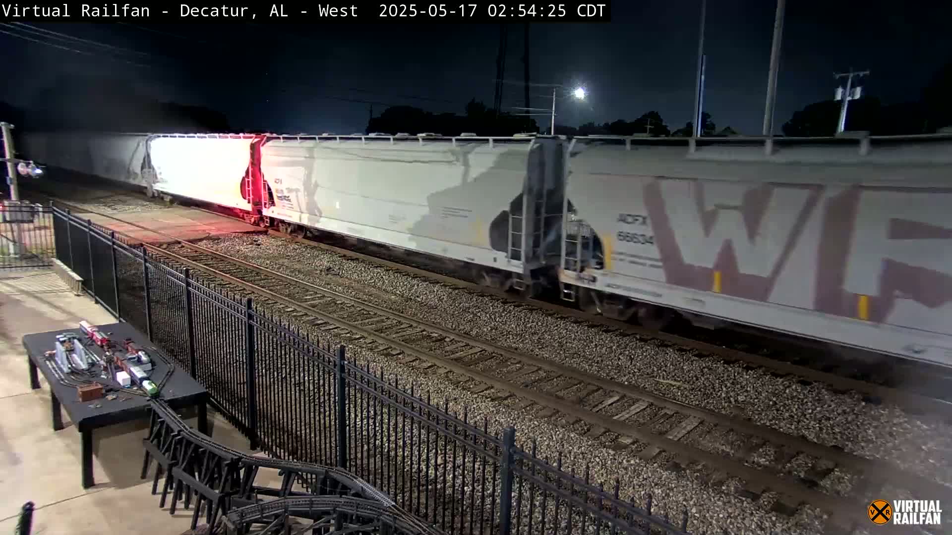 At night, a long freight train passes by a black metal fence, next to a model train set on a table.