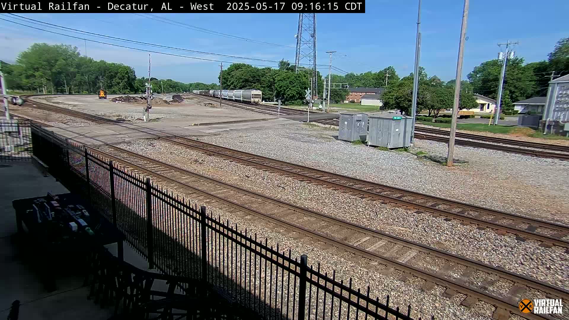 A sunny day at a railroad yard shows multiple train tracks, a few railcars, and some industrial buildings in the distance.