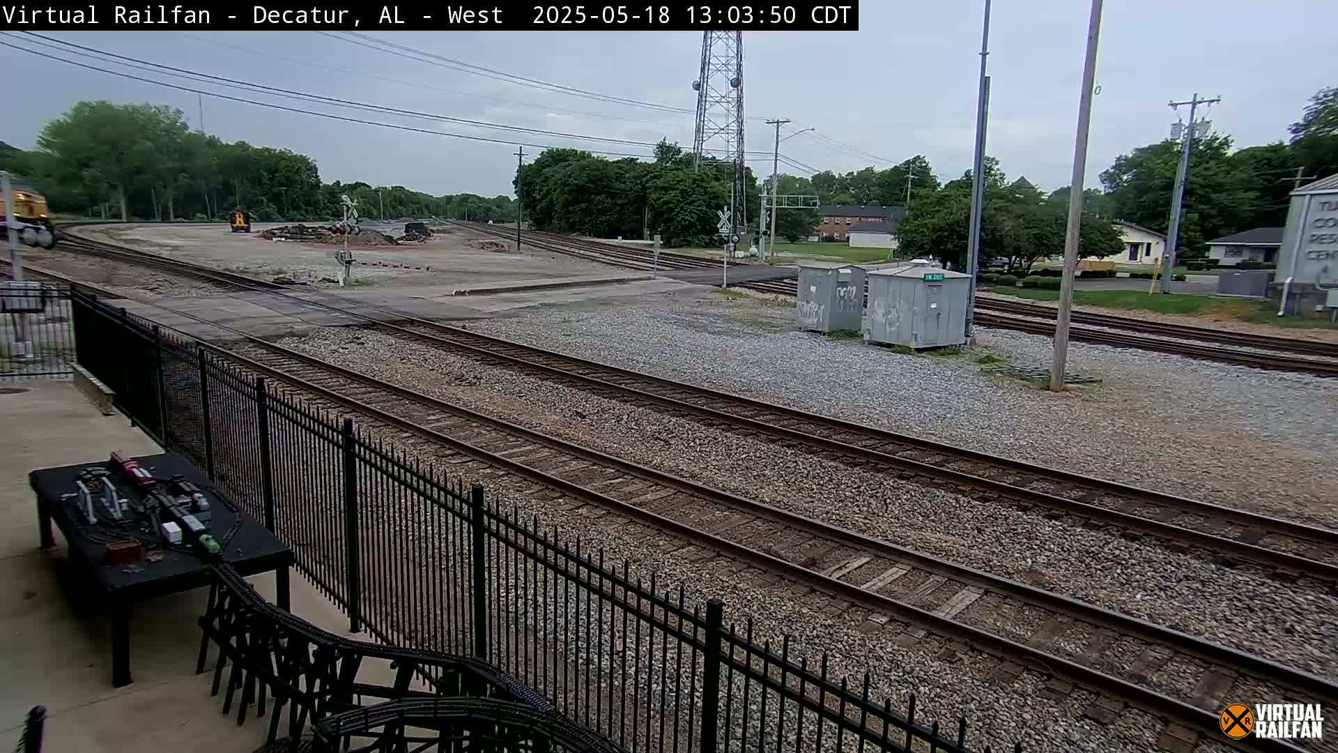 A partially cloudy day shows a railroad yard with several train tracks, a locomotive in the distance, and some buildings in the background.