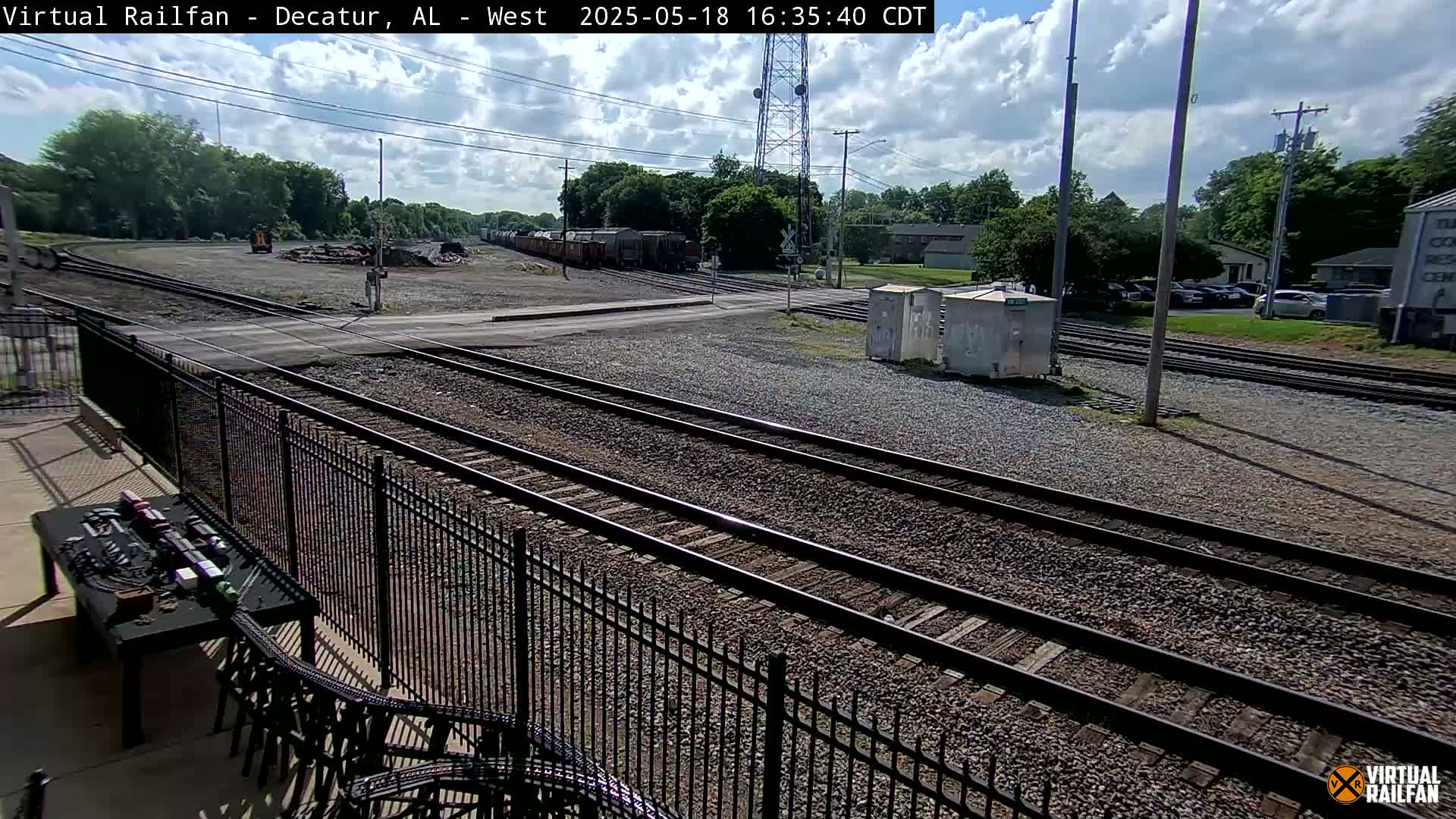 A sunny day shows multiple train tracks, a parked freight train, and a metal fence bordering a patio area with outdoor furniture.