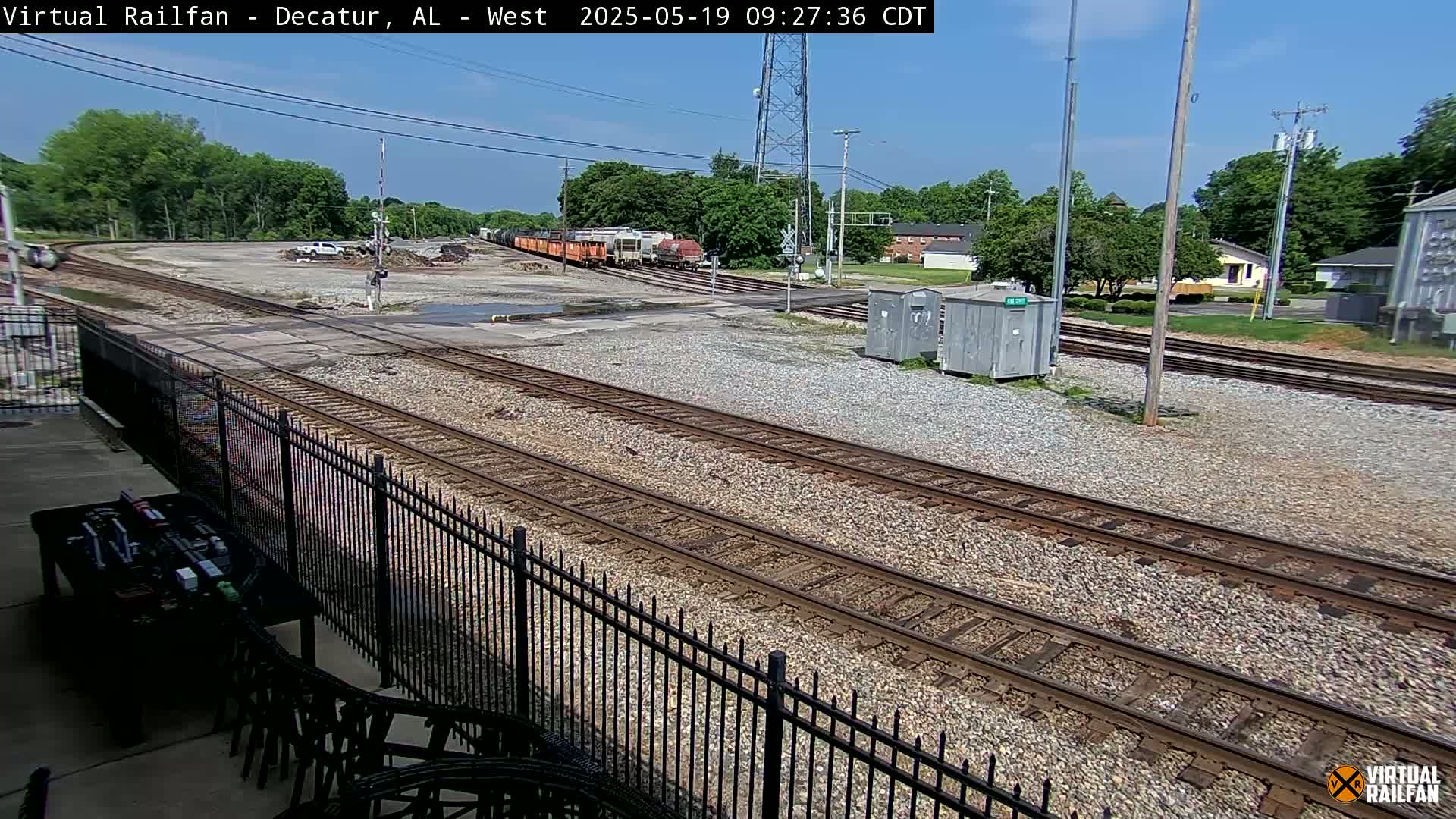 A sunny day reveals a railroad crossing with several train cars visible in the distance, bordered by a black metal fence and some buildings.