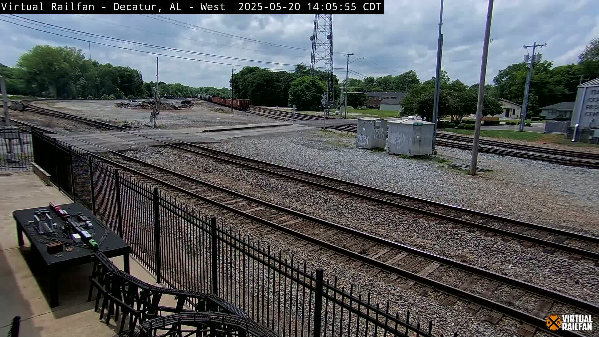 A view of railroad tracks and switching yards on a partly cloudy day, with trees and buildings in the background.
