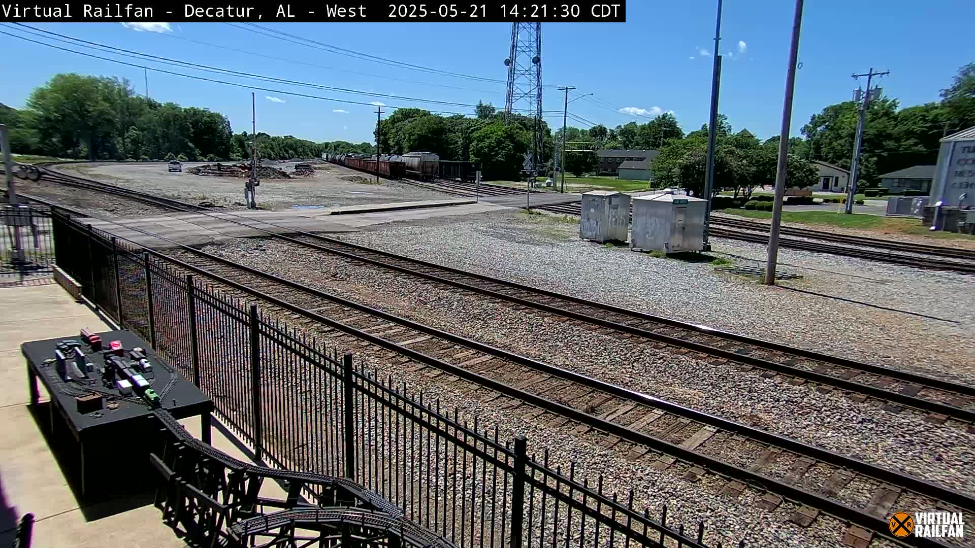 A sunny day view of several railway tracks intersecting near a fenced-in area, with a train car visible in the distance and a model train set nearby.