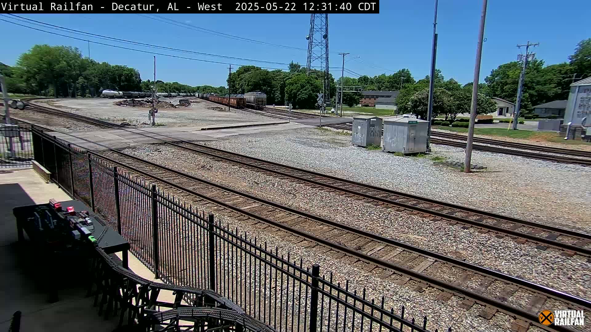 A sunny day reveals several sets of train tracks intersecting near a rail yard, with a black metal fence in the foreground.