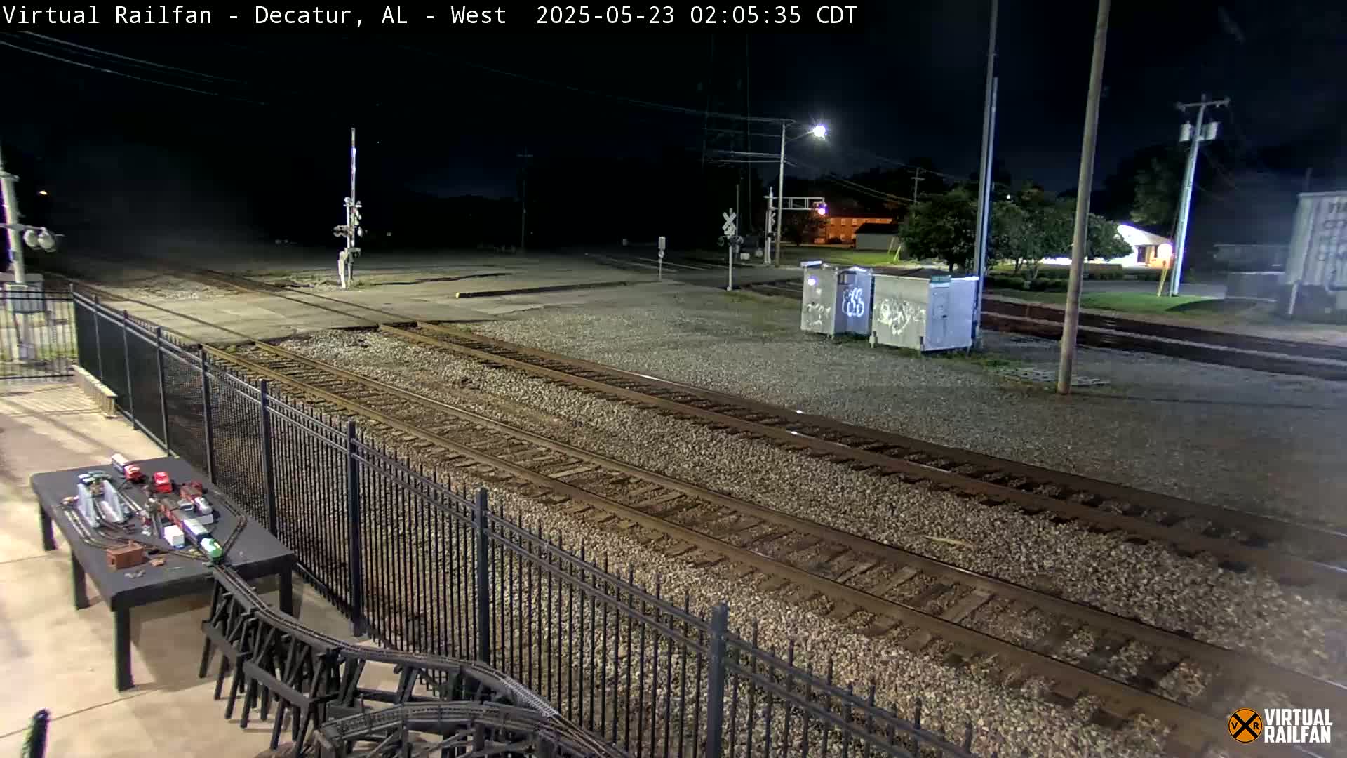 A nighttime view of railroad tracks, a metal fence, and a model train set on a table, under a dark sky with some lights visible in the distance.