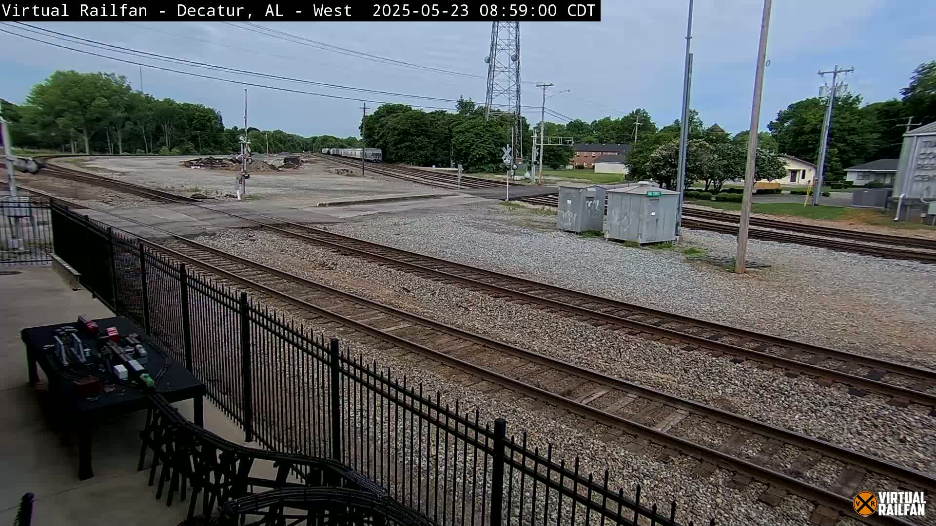 A view of a railroad yard with multiple train tracks, a black metal fence, and a partially visible building in the background under a partly cloudy sky.