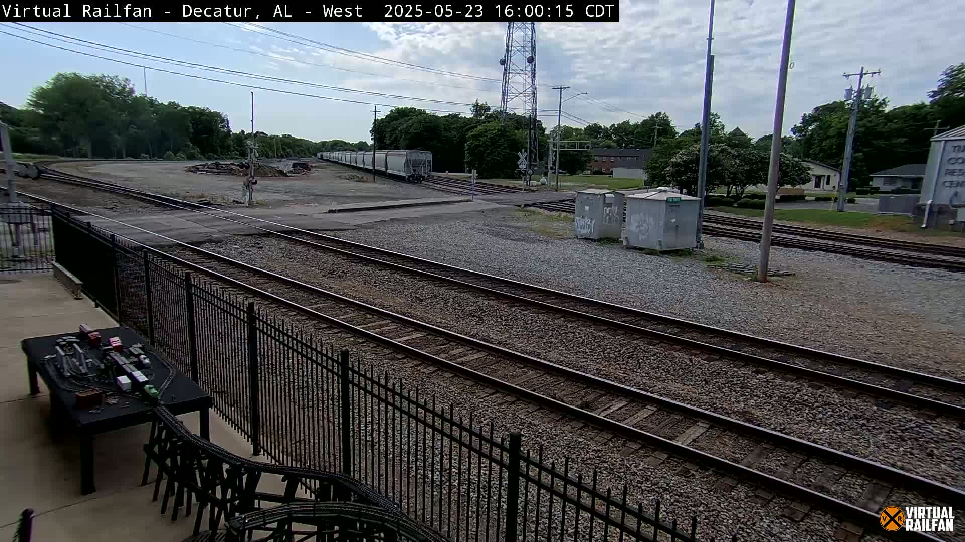 A mostly sunny view of a railway yard with multiple train tracks, a passing freight train, and surrounding trees and buildings.