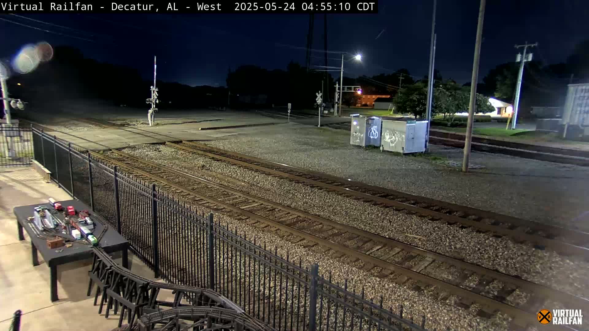 A nighttime view of railroad tracks, a metal fence, and a model train set on a table, under a clear, dark sky.