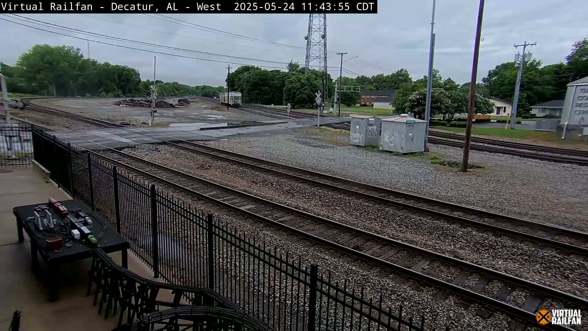 A railroad yard with several sets of tracks is visible on an overcast day, partially surrounded by a black metal fence, with a small building and some trees in the background.