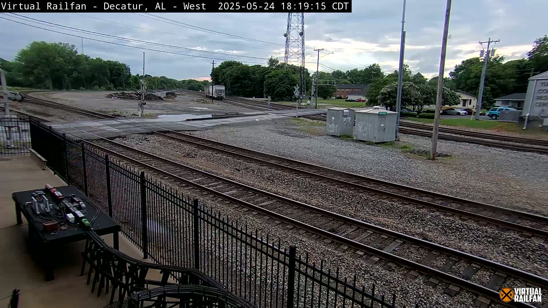 A view of railroad tracks and a street intersection under partly cloudy skies, with a fence and some buildings visible in the background.