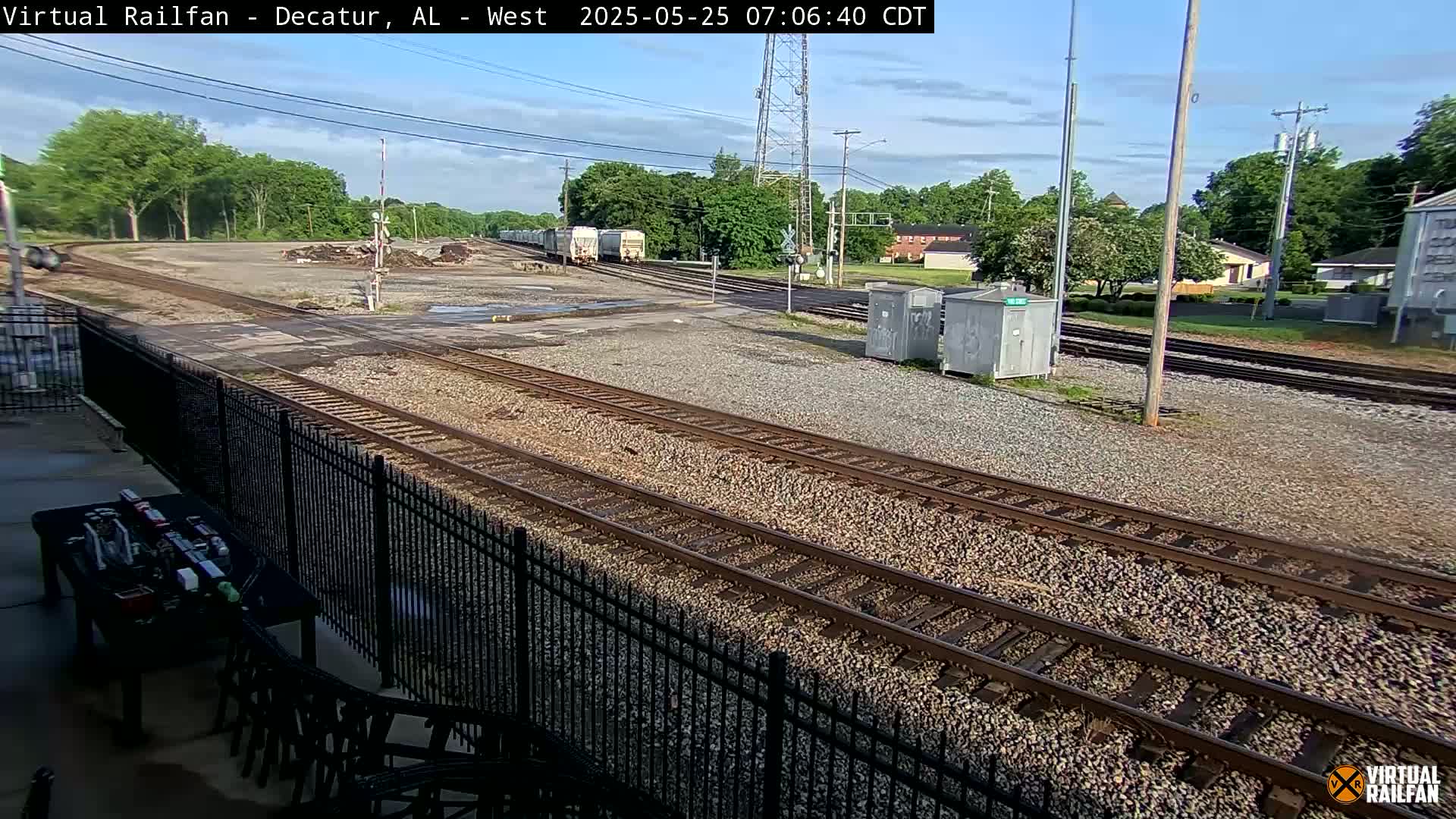 A mostly sunny outdoor scene shows several railroad tracks converging near a fenced-in area with a few boxcars visible in the distance.