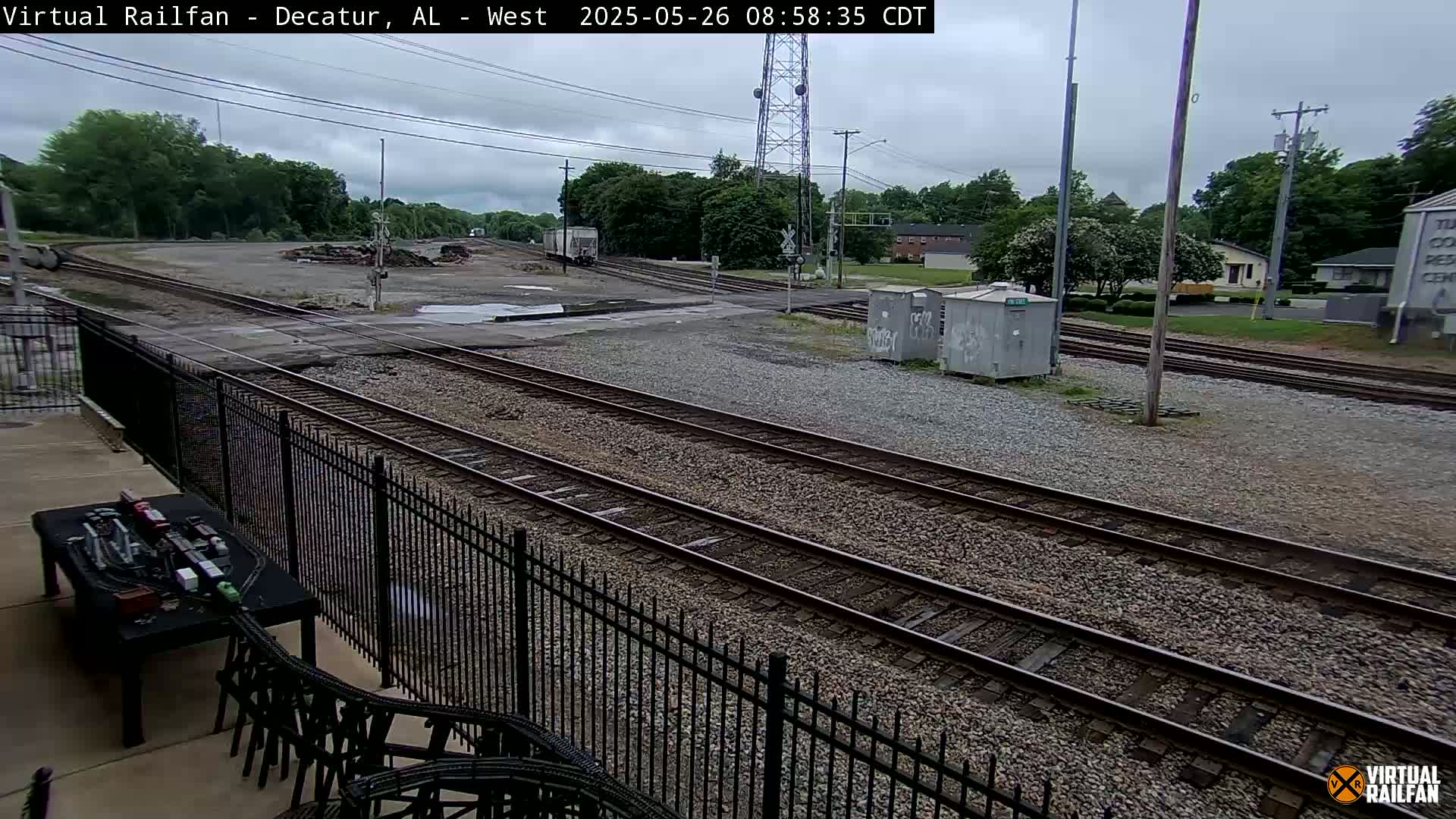 A view of railroad tracks and a gravel yard on an overcast day, seen from behind a black metal fence, with some buildings and trees in the background.