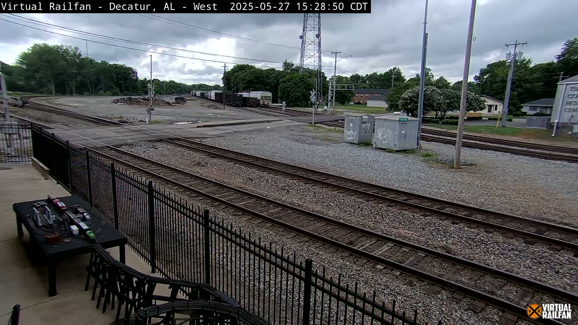 A railway junction with several sets of train tracks and a few railcars is visible under a partly cloudy sky.
