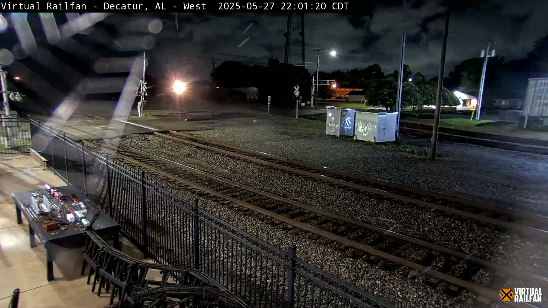 A nighttime view of railroad tracks under a cloudy sky, with a model train set visible on a table nearby.