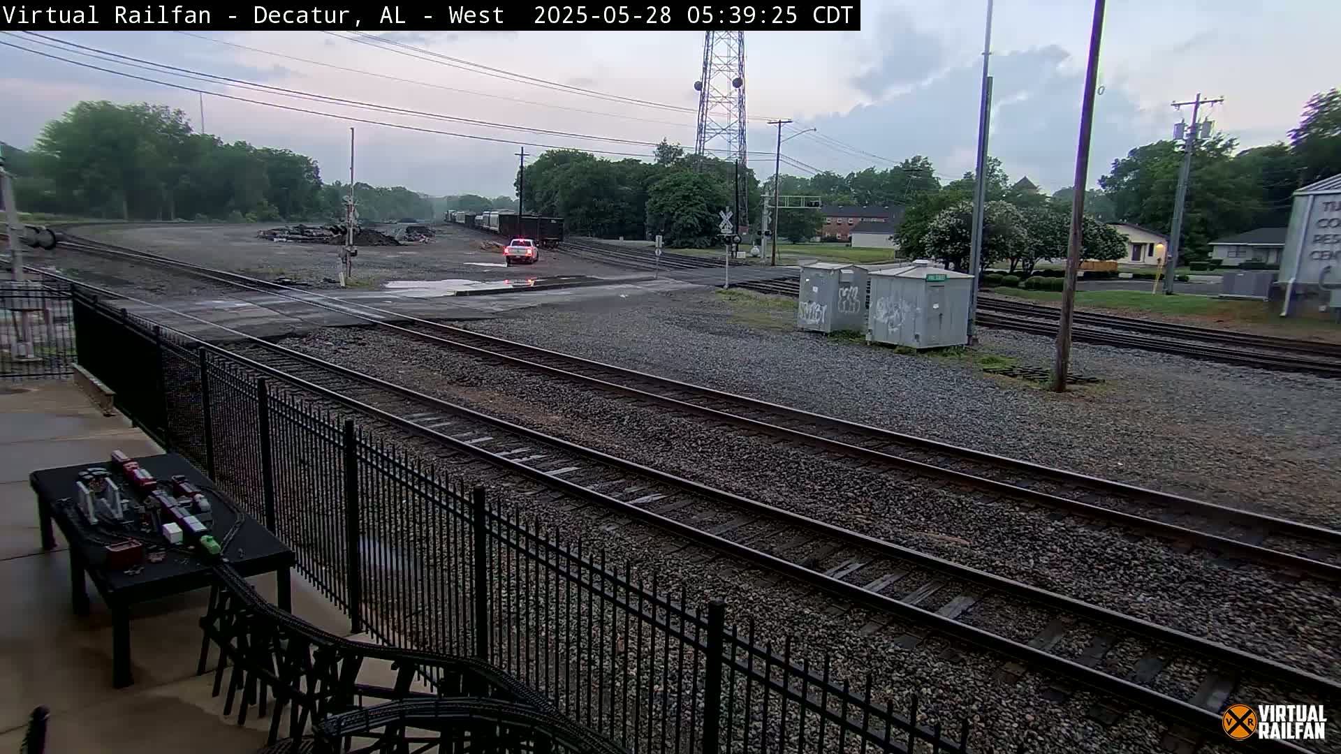 A view of a train yard with multiple train tracks, a vehicle, and surrounding buildings under an overcast sky.