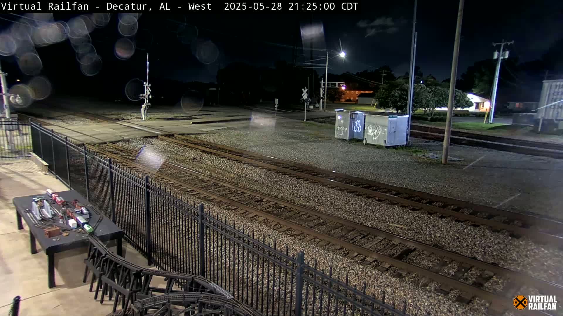 A nighttime view of train tracks, a model train set, and a black metal fence under a clear sky.