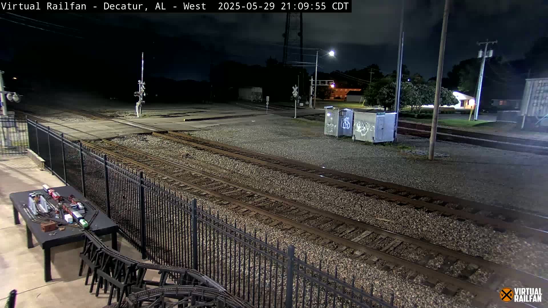 A nighttime view of a train yard with multiple sets of tracks, a black metal fence, and a model train set on a table in the foreground.