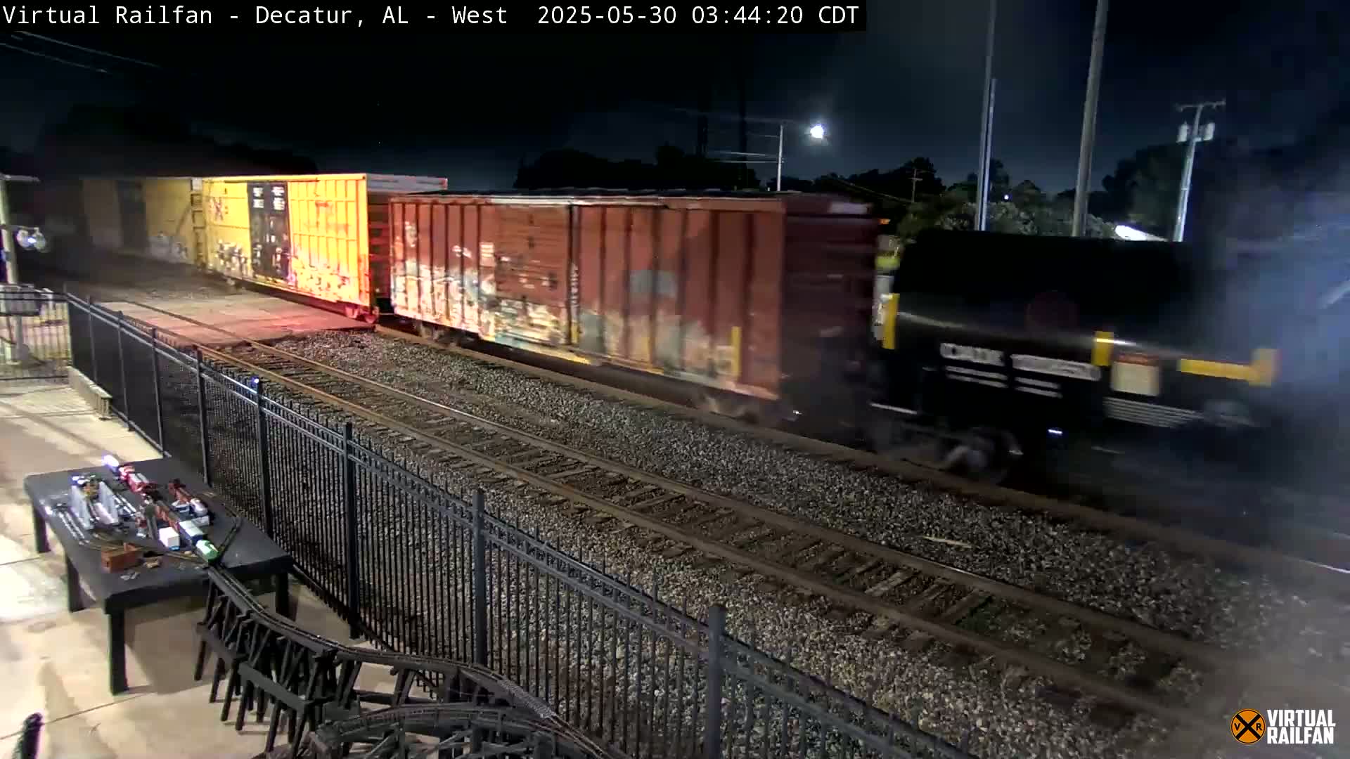 At night, a freight train with graffiti-covered cars passes by a black metal fence separating it from a table with a model train set.