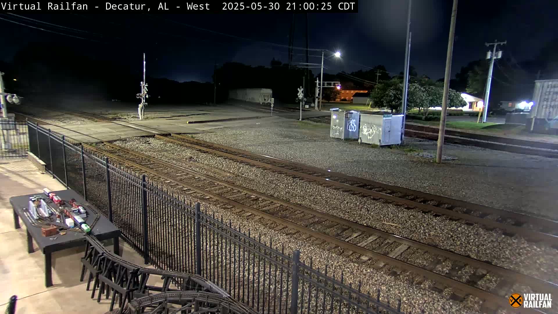 At night, a view of railroad tracks stretches into the distance, bordered by a black metal fence, with a model train set visible on a table in the foreground.